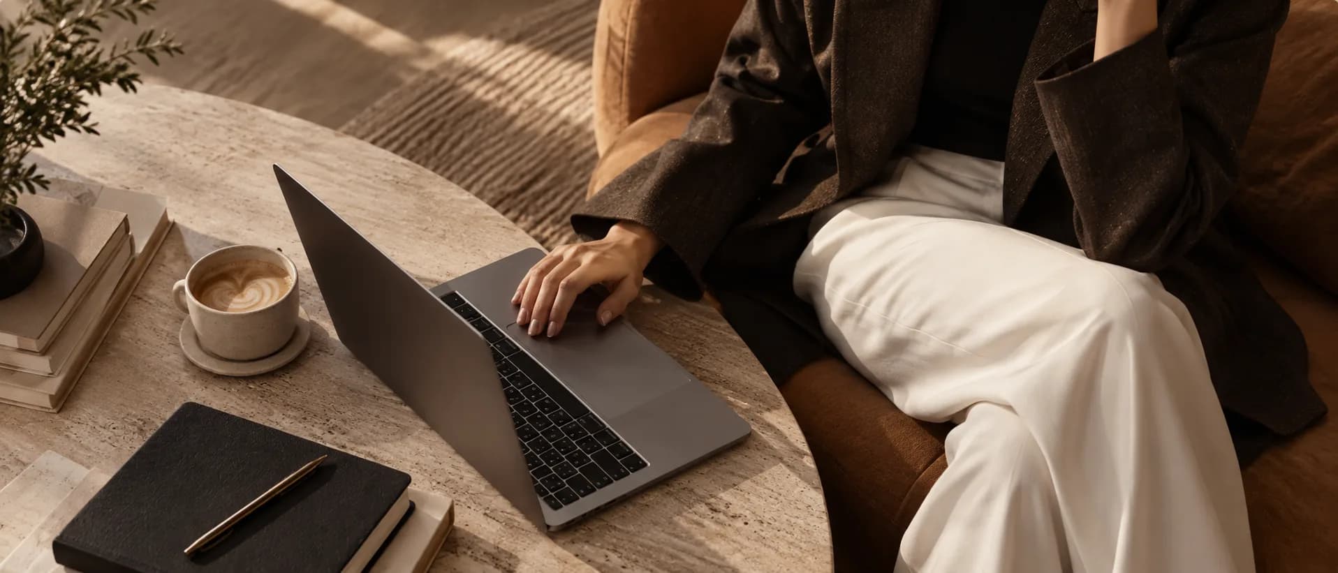 Someone in a dark blazer working on a laptop at a round travertine table in warm afternoon light, a coffee and a notebook beside it